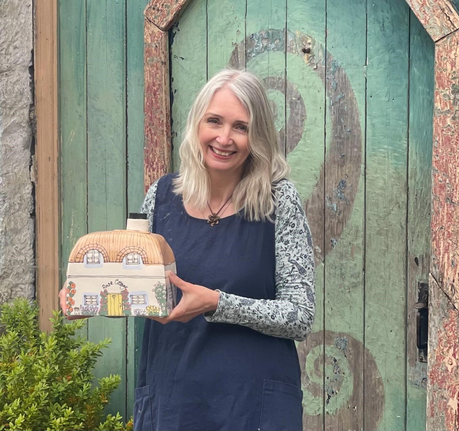 Potter Sam Hayes, blonde woman in blue dress, with ceramic urn painted like a country cottage.