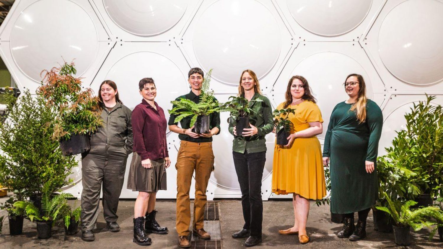 Six people , surrounded by plants and greenery, at Recompose's human composting facility in Seattle, behind them is an array of beehive shaped white metal vessels in which natural organic reduction takes place.