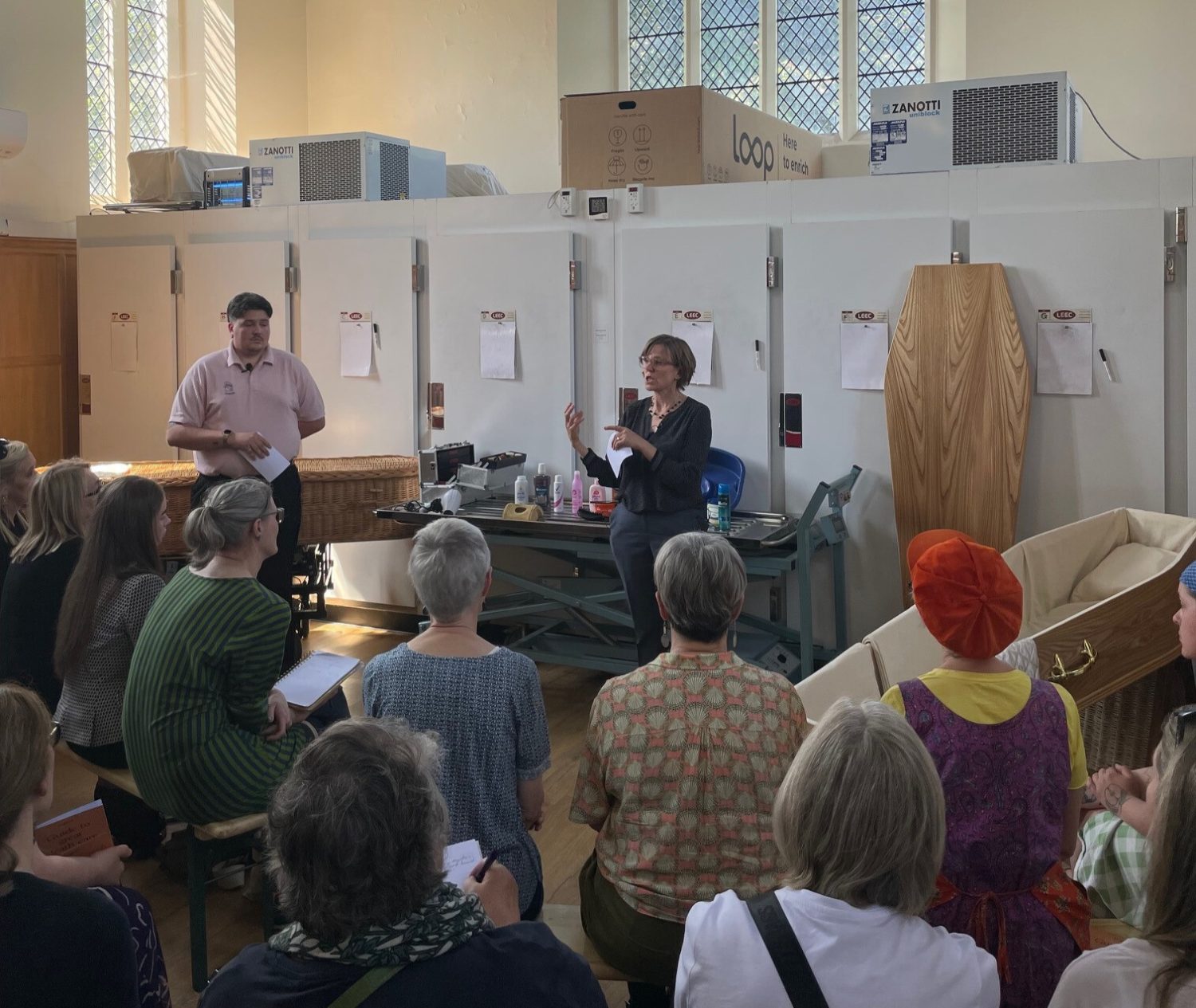 Liam and Hannah standing before a crowd in our mortuary talking. An empty calico lined coffin is on display beside Hannah