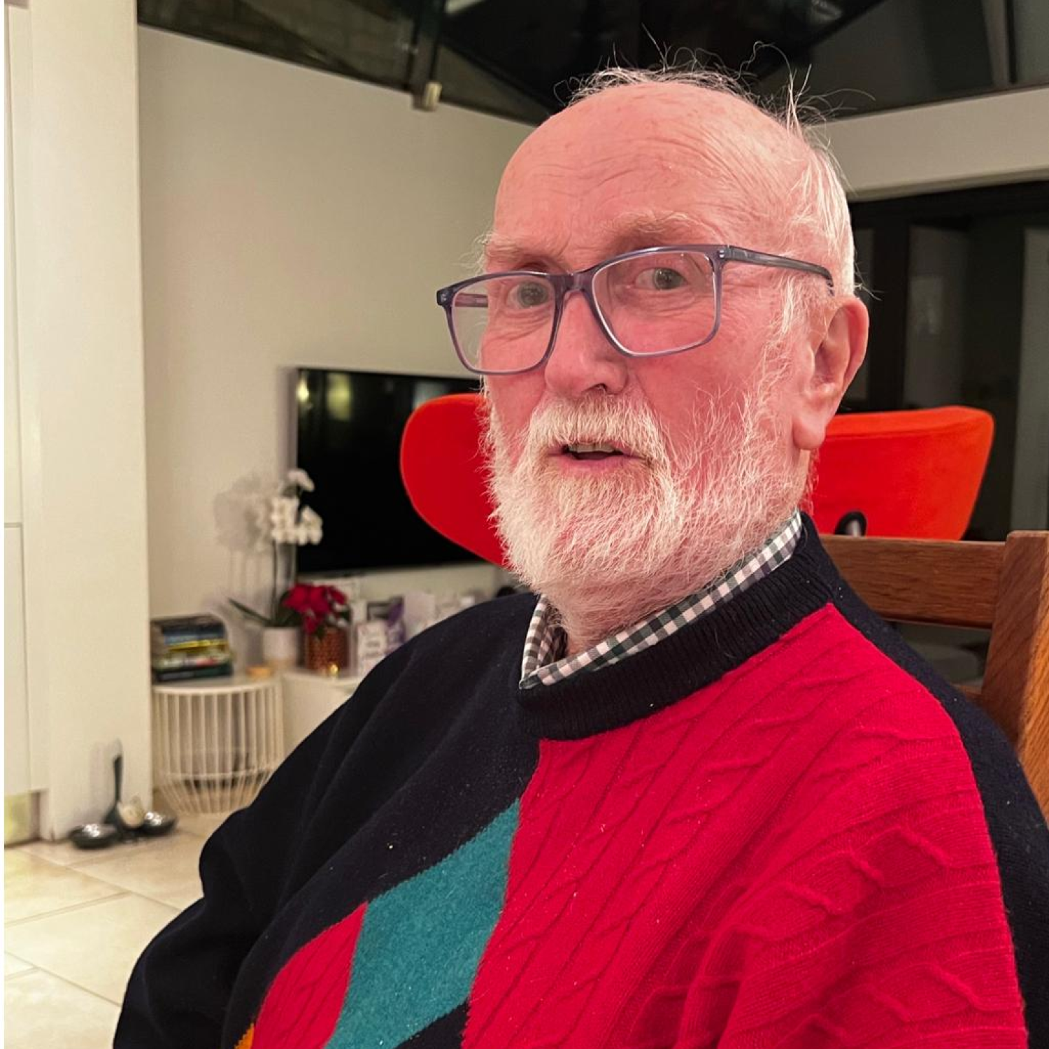 A man with glasses and a white beard, Derrick is wearing an argyle jumper and looking into the camera.
