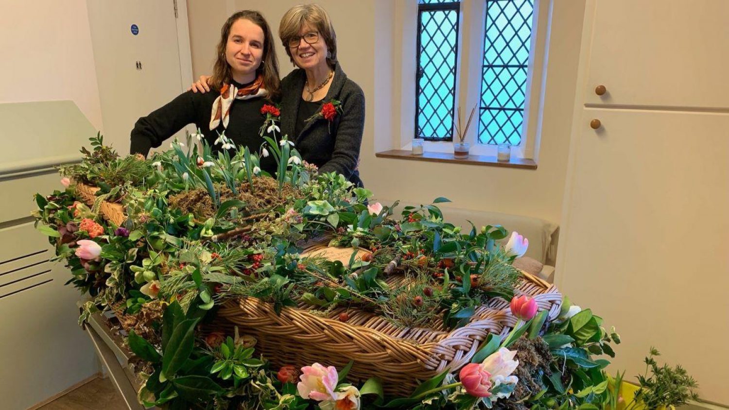 Two women next to willow coffin decorated with snowdrops and other flowers