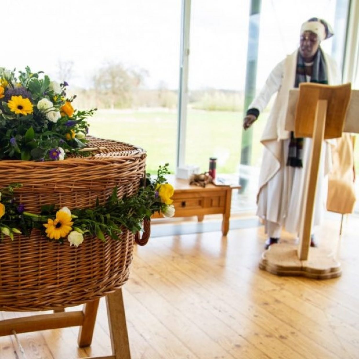 Willow coffin with yellow flowers, part-view of person wearing white, celebrant Aama Sade