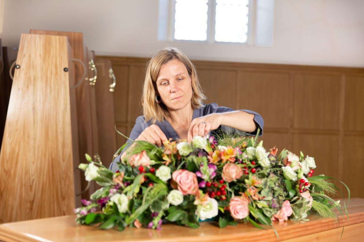 Woman arranging flowers on coffin, wooden coffins in background, at Poppy's, London ethical funeral director