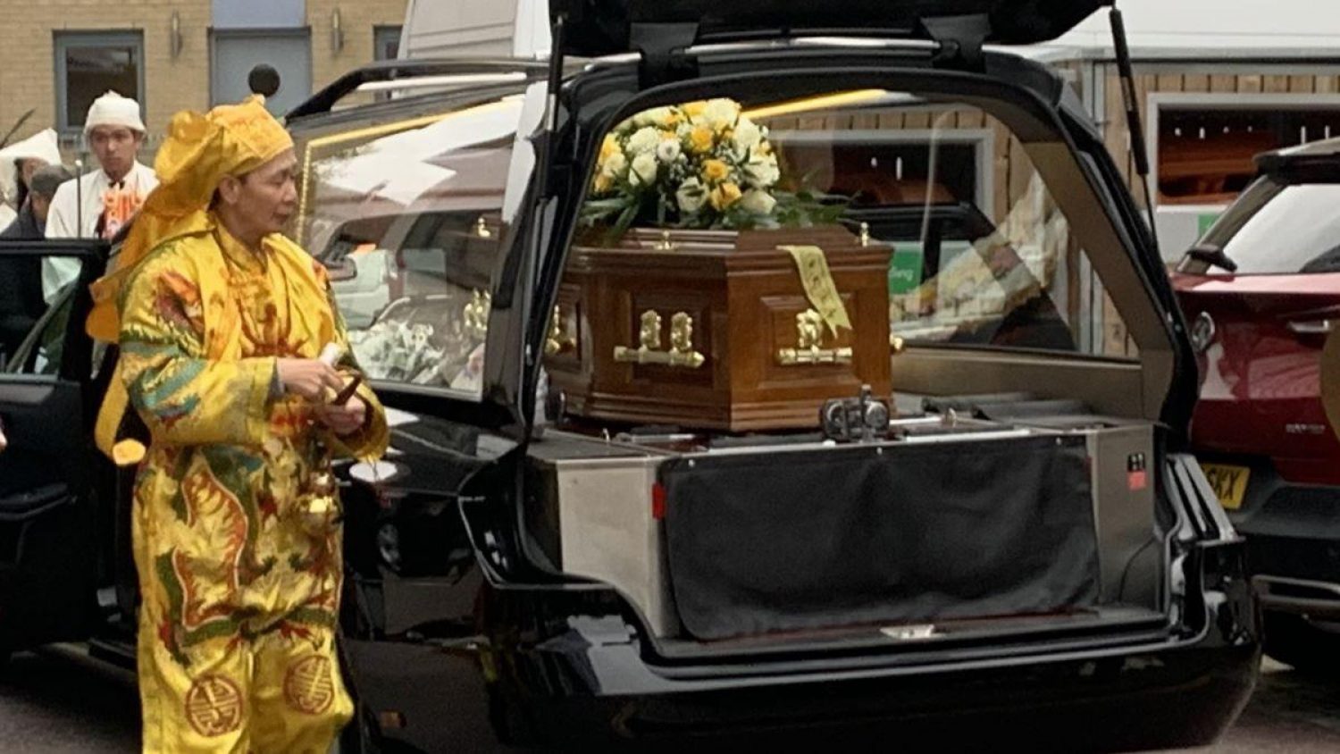 Taoist priest in yellow robes, next to coffin with yellow and white flowers in hearse, at Duc Lien's funeral