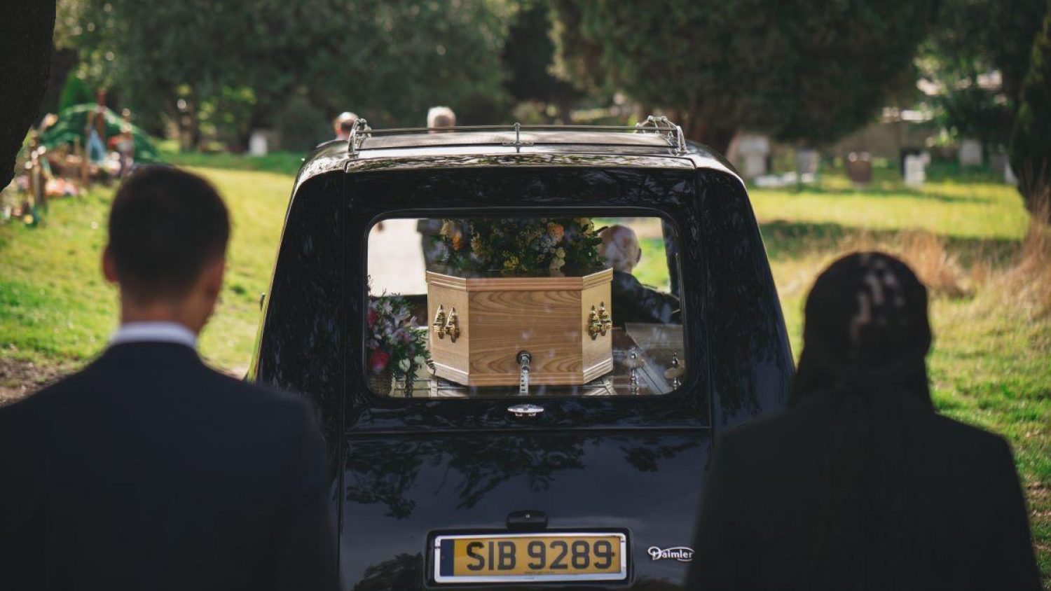 Two people following a black hearse with coffin in