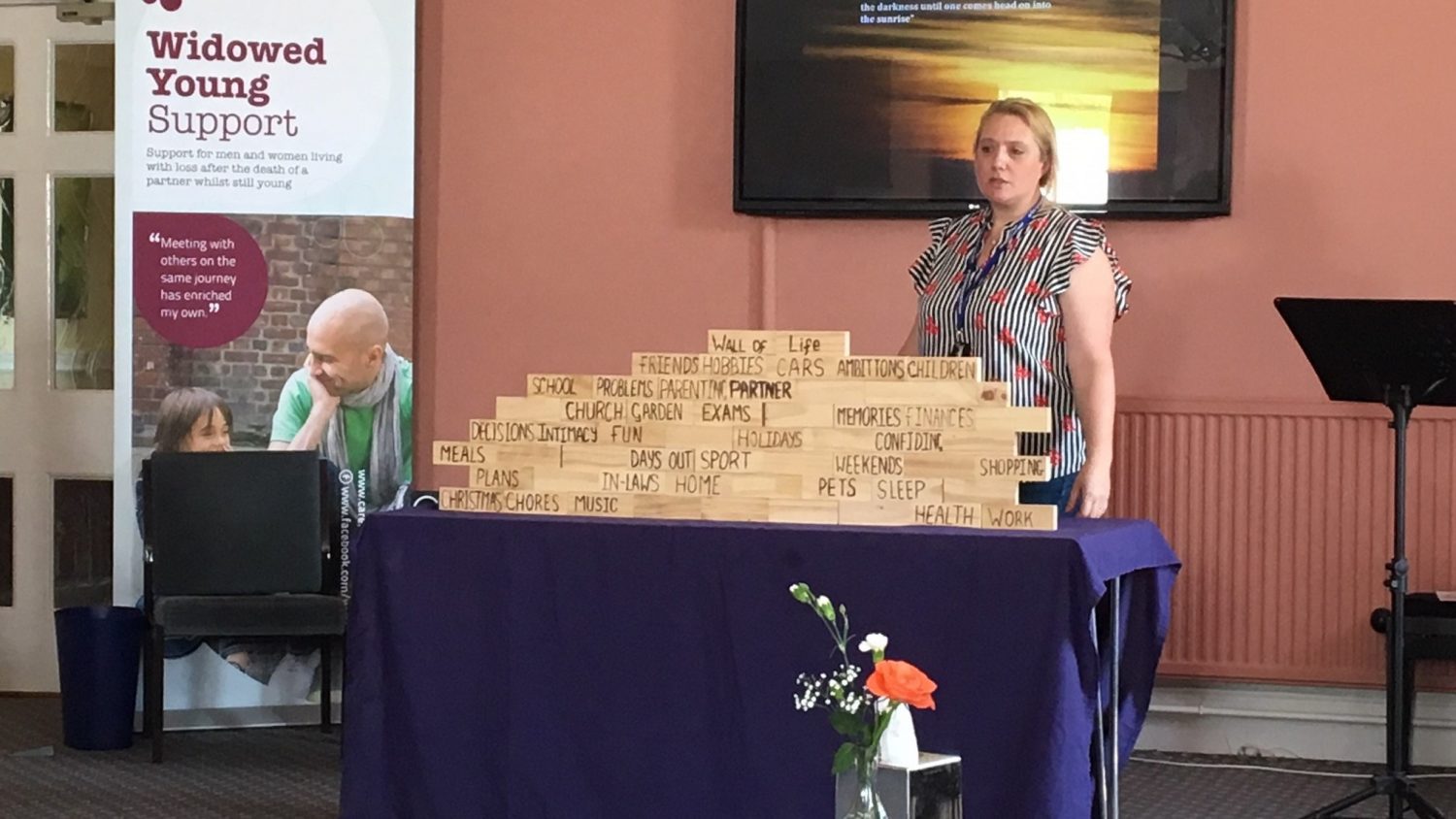 Facilitator at Widowed and Young support day, standing with building blocks to represent a wall of life (words like 'Christmas', 'children' and 'pets' are written on the blocks)