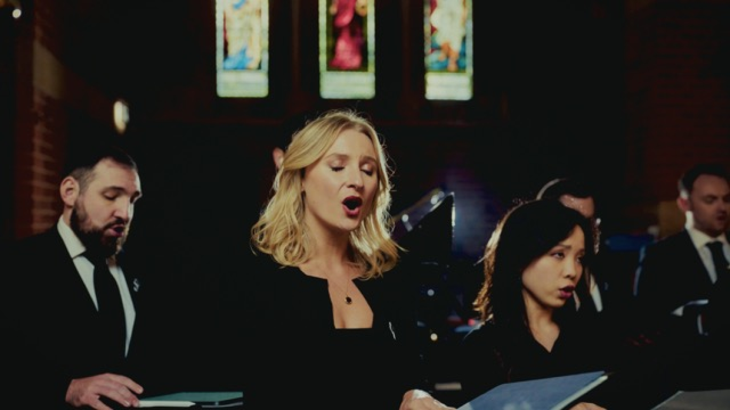 A blonde warm sings in the foreground, with other choir members behind her