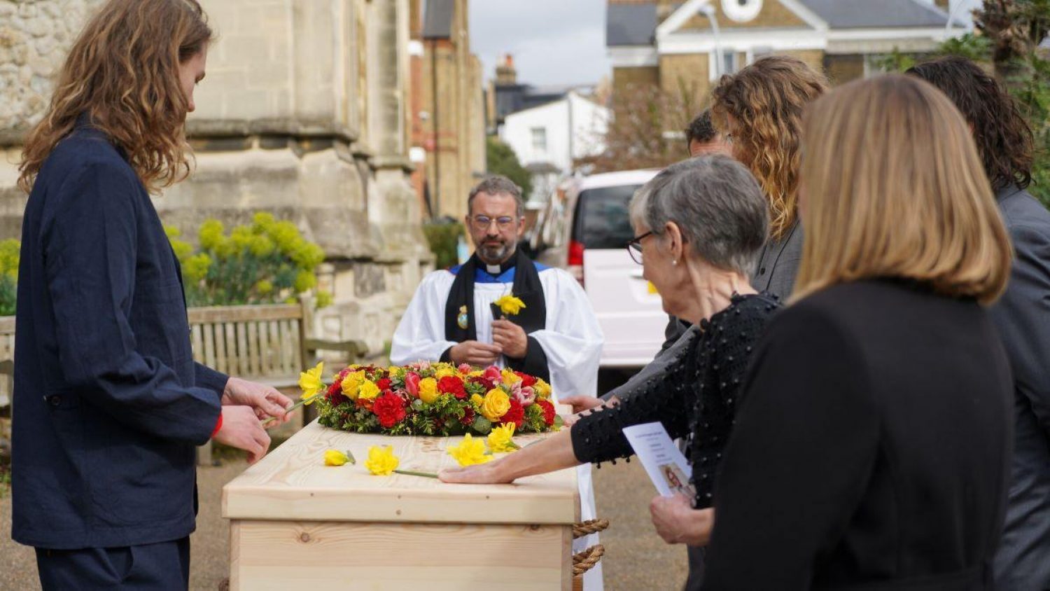 Vicar outside church with coffin, mourners placing daffodils on coffin