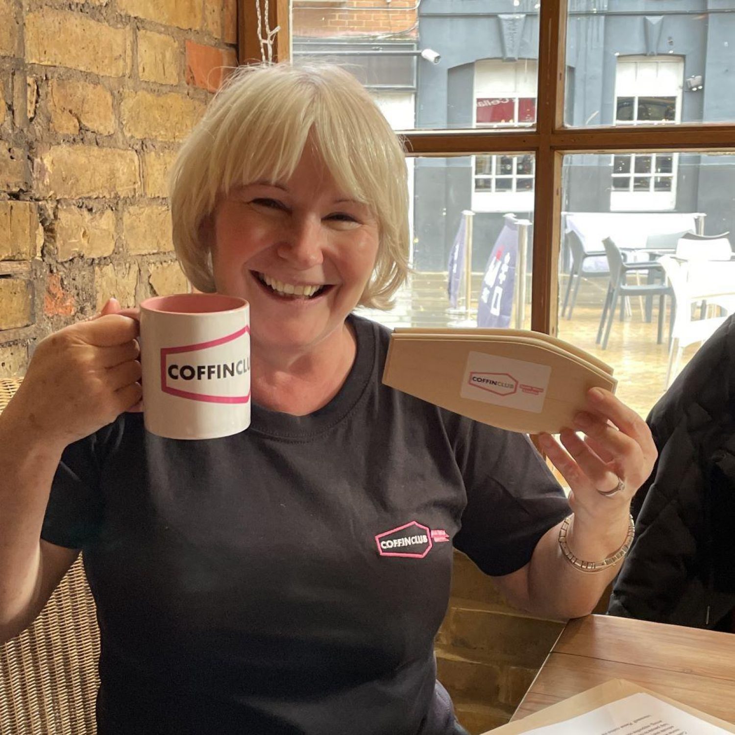White woman with short blond hair, holding coffin club branded mug and wearing coffin club t-shirt, smiling