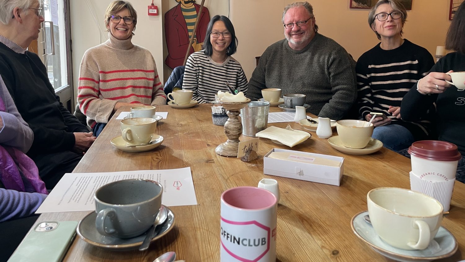Group of people sitting round table with mugs, coffin club branded mug in the foreground