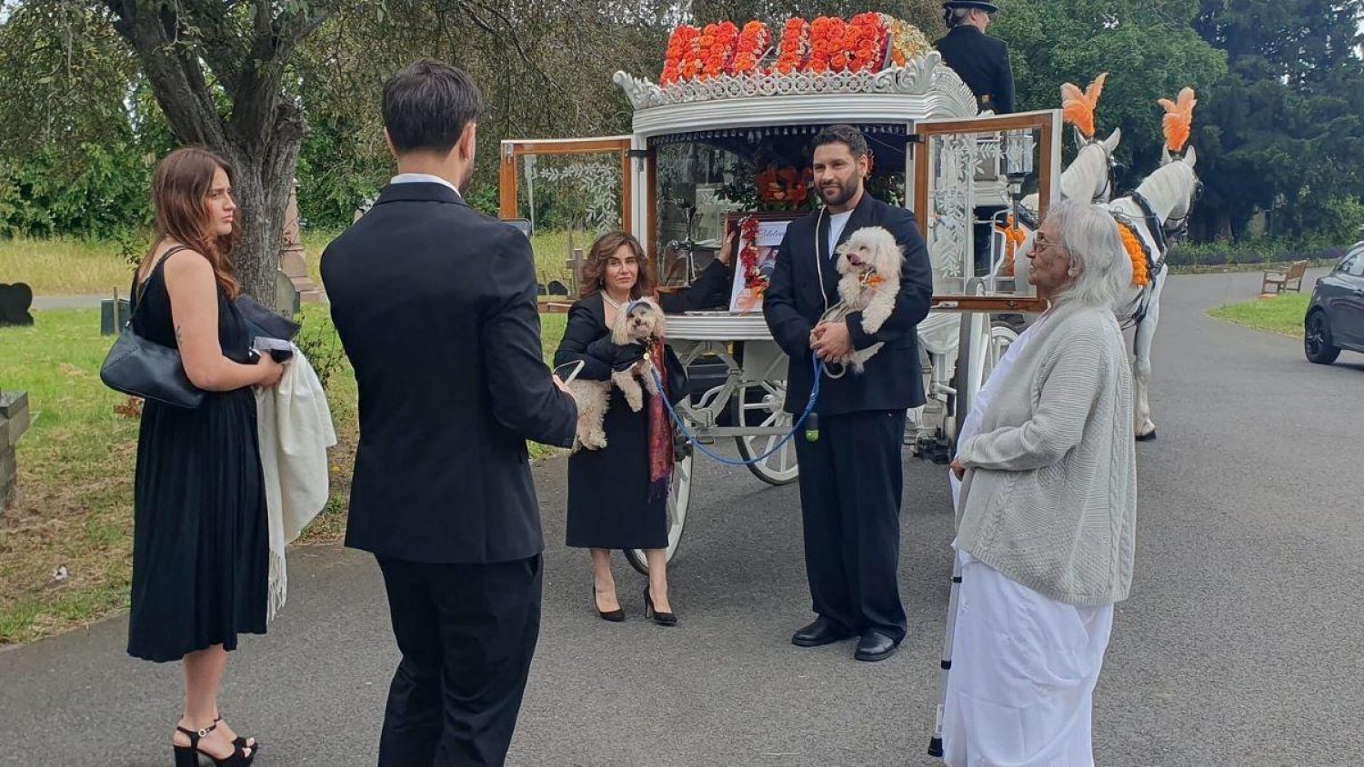 Family group next to white horse drawn hearse, two mourners are both holding dogs.