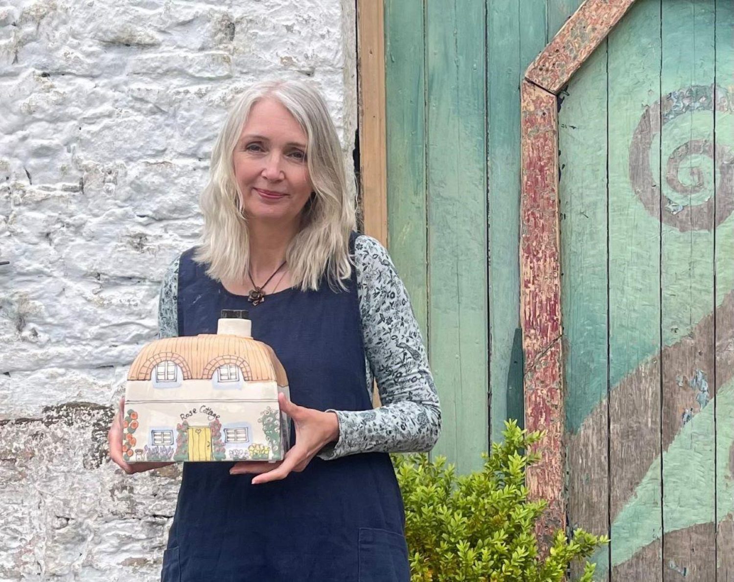 Potter Sam Hayes, woman with blonde hair and blue dress holding painted ceramic urn shaped like a country cottage
