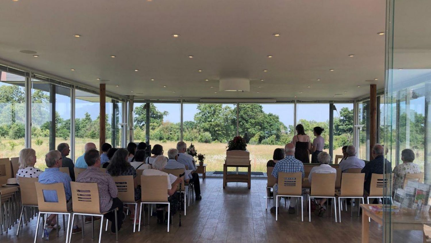 View of funeral congregation from back of chapel, large windows showing fields and sunshine
