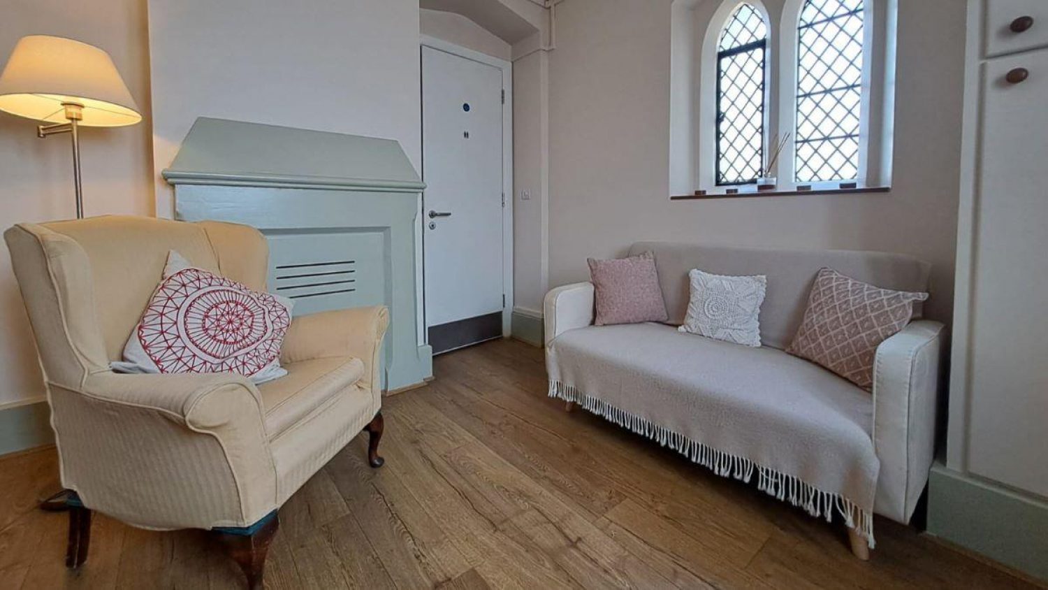 Room with sofa, armchair and standing lamp, light coloured walls and windows.