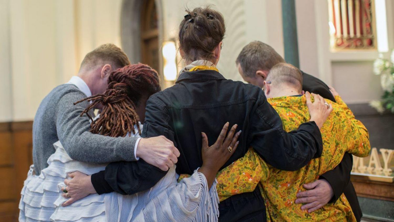Five people, different genders, ethnicities and ages with arms around each other in a crematorium