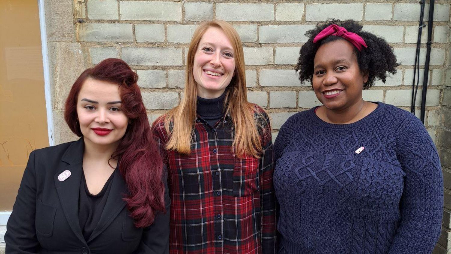 Three members of Poppy's team standing by Gatehouse office in Lambeth cemetery: Amy (white woman in black suit with red hair), Maia (white woman in checked shirt with long hair), Nichola (black woman in blue jumper with pink hair bow)